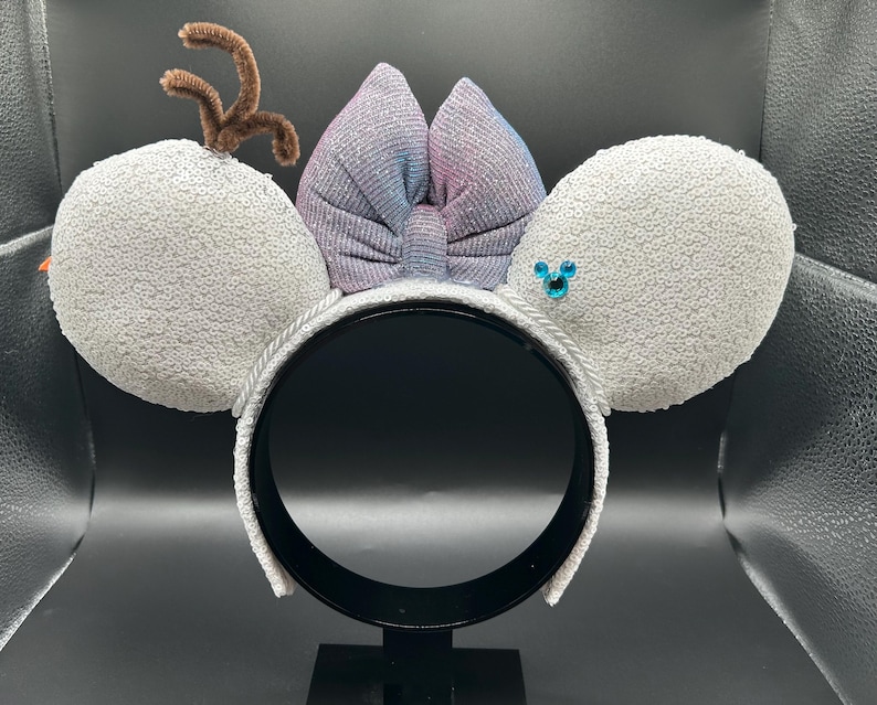 May include: A festive headband featuring large, sequined gray mouse ears. A shimmering bow in shades of purple and blue sits between the ears. Brown pipe cleaner antlers and a blue jewel accent complete the design. The headband is white.