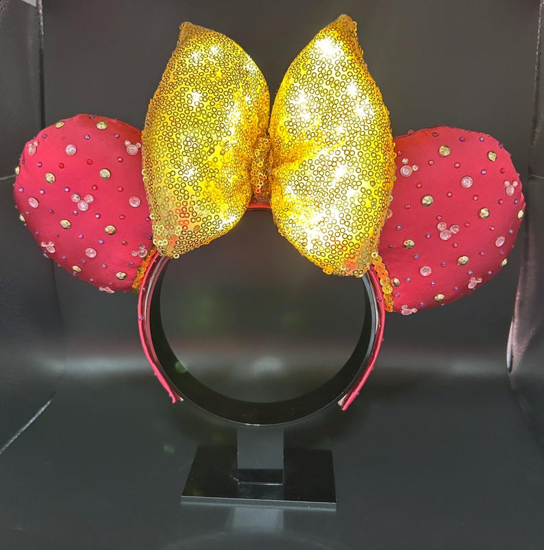 May include: A pair of mouse ears with a gold sequined bow and red ears. The red ears are embellished with small, clear rhinestones. The headband is black with a red trim. The ears are on a black stand.