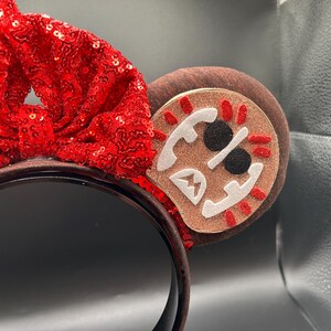May include: A headband featuring a large, bright red sequined bow centered between two brown ears. The right ear displays a circular patch with a glittery tan background, white felt accents, black circular eyes, and red stitched details.