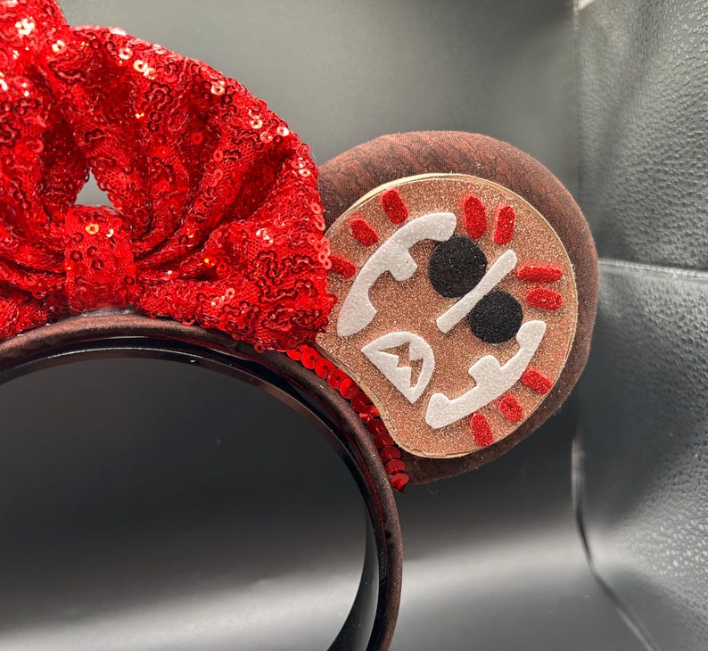 May include: A headband featuring a large, bright red sequined bow centered between two brown ears. The right ear displays a circular patch with a glittery tan background, white felt accents, black circular eyes, and red stitched details.
