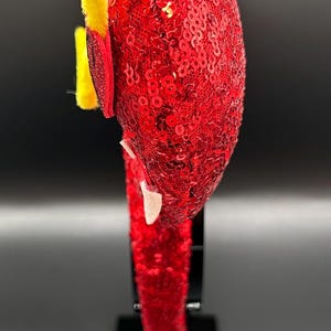 May include: A red sequined headband with yellow pipe cleaner accents. The headband is decorated with red and white embellishments. The item is displayed on a black stand against a dark background.