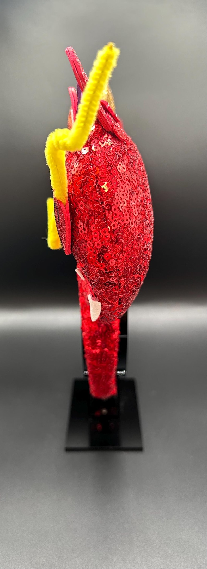 May include: A red sequined headband with yellow pipe cleaner accents. The headband is decorated with red and white embellishments. The item is displayed on a black stand against a dark background.