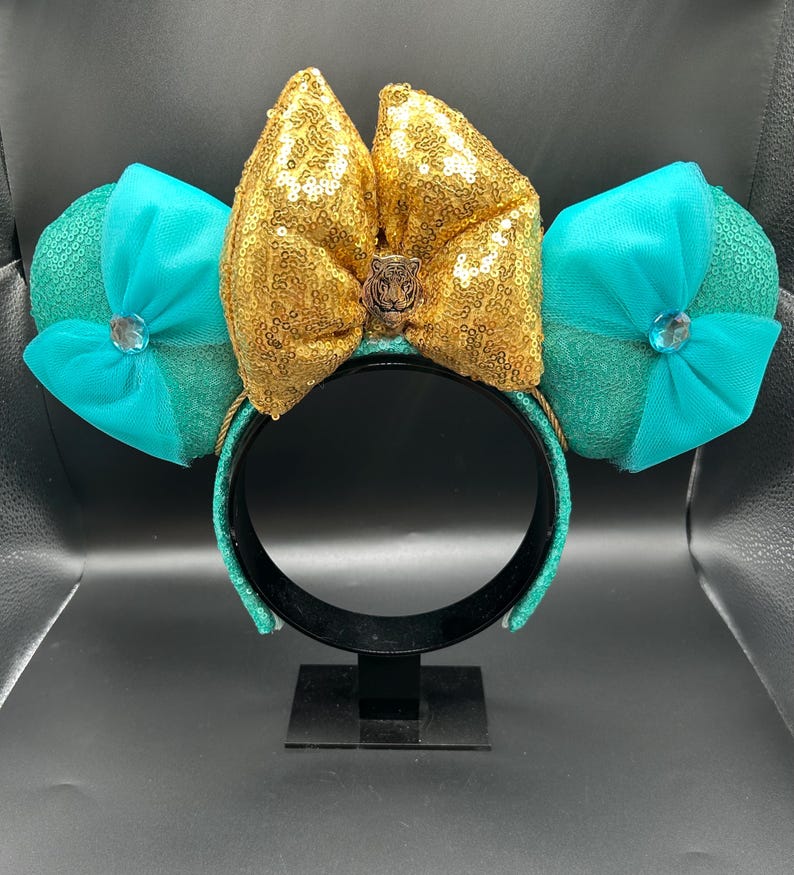 May include: A teal and gold sequined headband with large ears. The ears are teal with a bow and a jewel. The centre bow is gold with a tiger emblem. The headband is on a black stand.