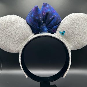May include: A pair of mouse ears with white sequined ear pads and a dark blue bow with a galaxy print. The headband is white with a black interior. A small blue gem is on one ear.
