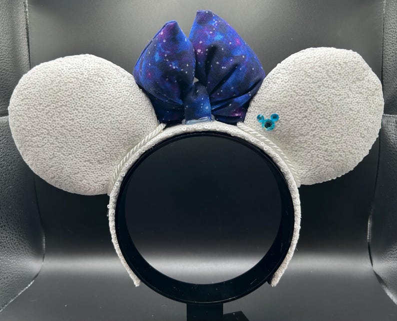 May include: A pair of mouse ears with white sequined ear pads and a dark blue bow with a galaxy print. The headband is white with a black interior. A small blue gem is on one ear.
