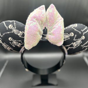 May include: Black and white skeleton-themed mouse ears with a sequined bow. The ears are black with a repeating skeleton pattern. The bow is light pink with iridescent sequins. The headband is black.