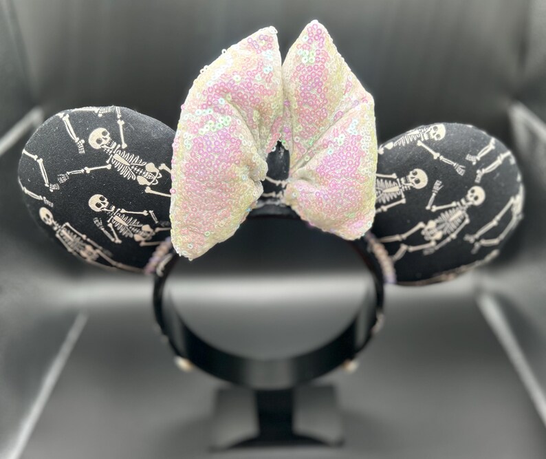 May include: Black and white skeleton-themed mouse ears with a sequined bow. The ears are black with a repeating skeleton pattern. The bow is light pink with iridescent sequins. The headband is black.