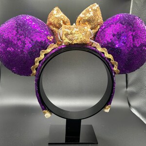 Pu&ograve; includere: Orecchie di Minnie Mouse con paillettes viola e dorate con un fiocco dorato. Le orecchie sono su un supporto nero.