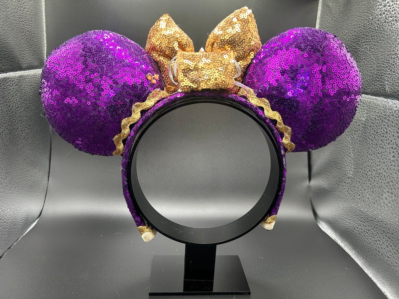 Pu&ograve; includere: Orecchie di Minnie Mouse con paillettes viola e dorate con un fiocco dorato. Le orecchie sono su un supporto nero.