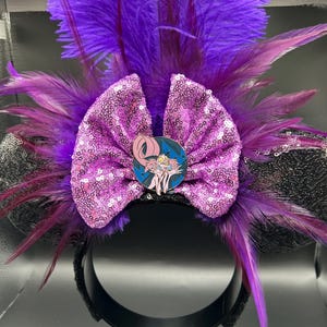 May include: A headband featuring black ears, a large pink sequined bow, and vibrant purple feathers. A circular enamel pin depicting a pink, long-tailed creature with yellow eyes and sharp teeth is centered on the bow, set against a dark blue background.