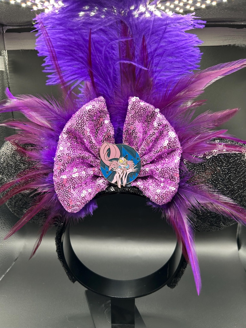 May include: A headband featuring black ears, a large pink sequined bow, and vibrant purple feathers. A circular enamel pin depicting a pink, long-tailed creature with yellow eyes and sharp teeth is centered on the bow, set against a dark blue background.