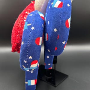 K&ouml;nnte beinhalten: Ein patriotisches Stirnband mit einem roten Paillettenakzent, einem wei&szlig;en Paillettenakzent und zwei blauen Stoffst&uuml;cken. Der blaue Stoff ist mit wei&szlig;en Sternen und Eis am Stiel-Grafiken gemustert. Das Stirnband wird auf einem schwarzen St&auml;nder ausgestellt.