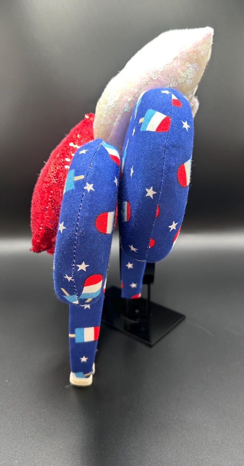 K&ouml;nnte beinhalten: Ein patriotisches Stirnband mit einem roten Paillettenakzent, einem wei&szlig;en Paillettenakzent und zwei blauen Stoffst&uuml;cken. Der blaue Stoff ist mit wei&szlig;en Sternen und Eis am Stiel-Grafiken gemustert. Das Stirnband wird auf einem schwarzen St&auml;nder ausgestellt.