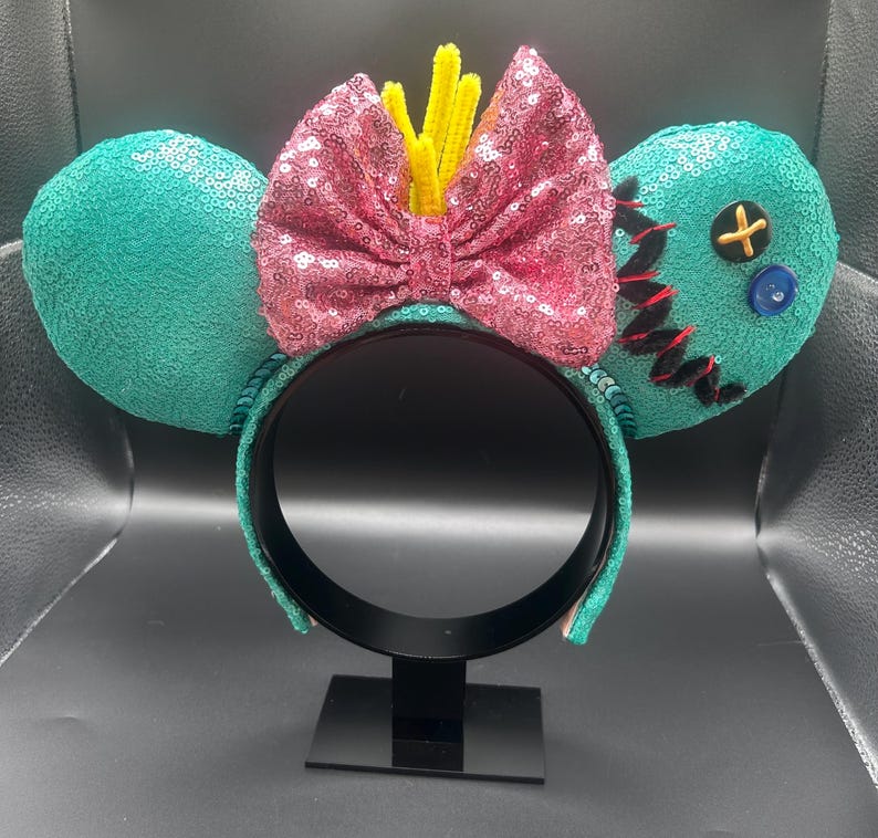 May include: A teal sequined headband with two round ears. A pink sequined bow with yellow accents sits between the ears. One ear has black stitching and a yellow cross button. A blue button is also present.