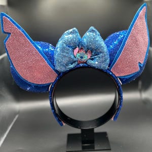 May include: A blue sequined headband with large, pink glittered ears and a matching bow. The bow features a small Stitch character emblem. The headband is a fun accessory for themed events or cosplay.