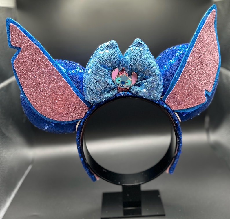 May include: A blue sequined headband with large, pink glittered ears and a matching bow. The bow features a small Stitch character emblem. The headband is a fun accessory for themed events or cosplay.