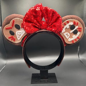 May include: A brown headband featuring two circular ears with gingerbread-inspired faces. Each ear has a light brown glittery center with white, black, and red felt details. A large, bright red sequined bow sits between the two ears.