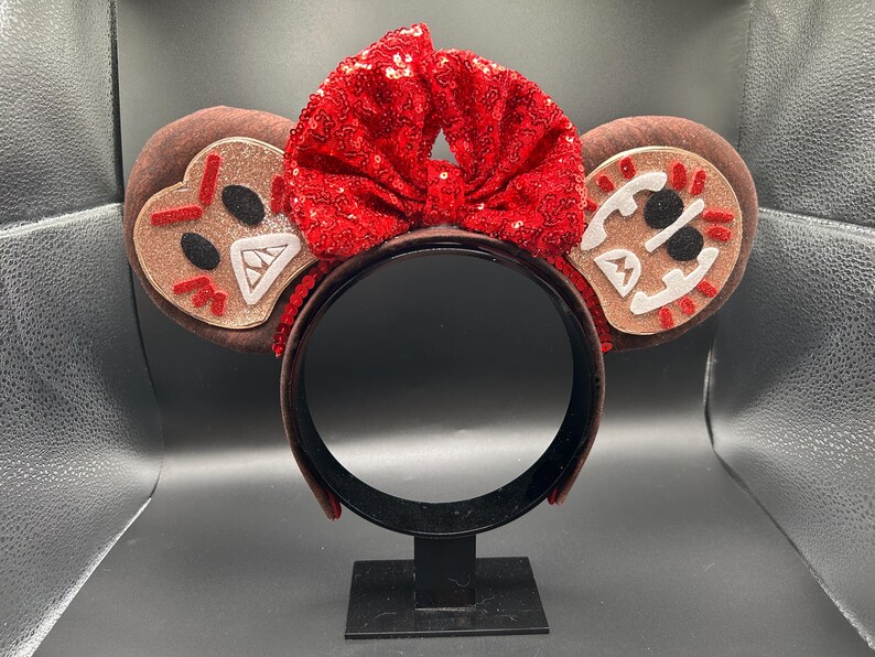 May include: A brown headband featuring two circular ears with gingerbread-inspired faces. Each ear has a light brown glittery center with white, black, and red felt details. A large, bright red sequined bow sits between the two ears.