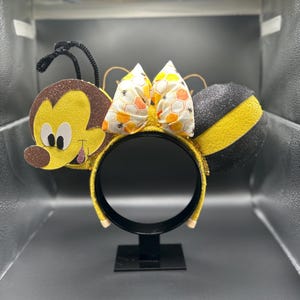 May include: A pair of yellow and black bee-themed Minnie Mouse ears with a honeycomb bow. The ears are made of felt and have a glitter finish.