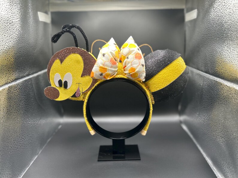 May include: A pair of yellow and black bee-themed Minnie Mouse ears with a honeycomb bow. The ears are made of felt and have a glitter finish.