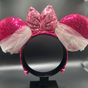 Op de afbeelding: Roze glitzerende Minnie Mouse oren haarband met een grote roze strik en witte tule.