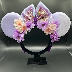 Puede incluir: Una diadema con dos grandes orejas circulares de color morado claro y un lazo central de tela iridiscente con estampado floral. La diadema est&aacute; decorada con un arreglo de flores artificiales en tonos morado claro y crema.
