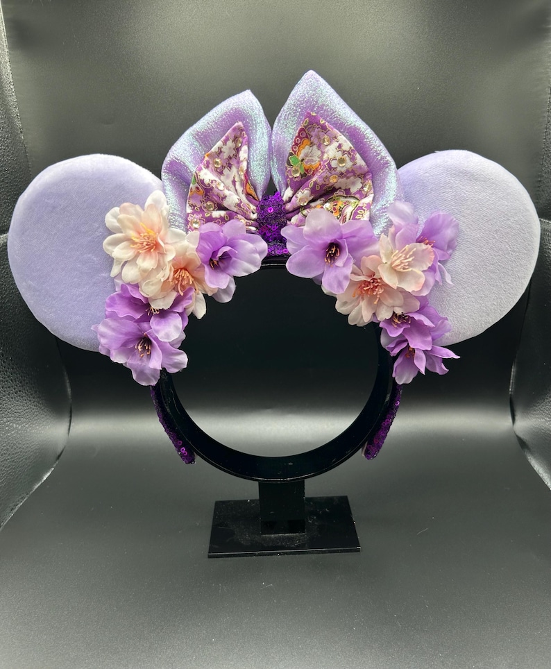 Puede incluir: Una diadema con dos grandes orejas circulares de color morado claro y un lazo central de tela iridiscente con estampado floral. La diadema est&aacute; decorada con un arreglo de flores artificiales en tonos morado claro y crema.