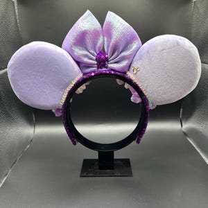 Puede incluir: Una diadema con dos orejas grandes y redondas de terciopelo morado claro y un lazo central morado iridiscente. La estructura de la diadema est&aacute; cubierta de lentejuelas moradas oscuras, con un peque&ntilde;o detalle plateado de Mickey Mouse en la oreja derecha.