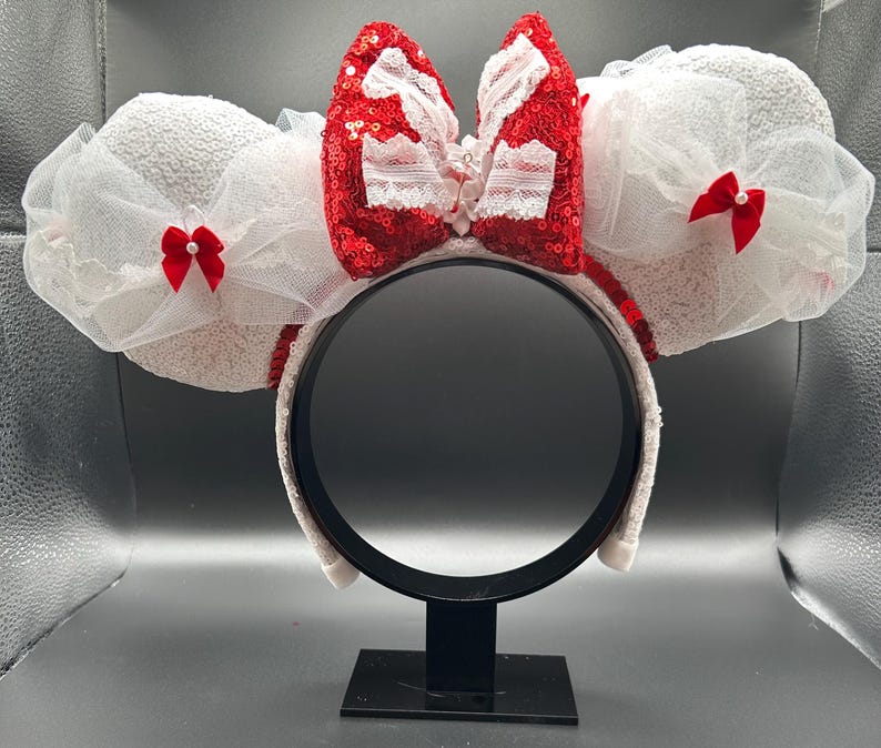 May include: White Minnie Mouse ears with a large red sequined bow and white lace trim. The bow has a small red bow on each side.