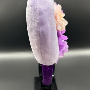 Puede incluir: Una vista lateral de una diadema con una oreja circular de felpa de color morado claro. La oreja est&aacute; decorada con flores artificiales en capas en tonos rosa p&aacute;lido y morado oscuro. La base de la diadema est&aacute; cubierta con un tejido texturizado de color morado oscuro.