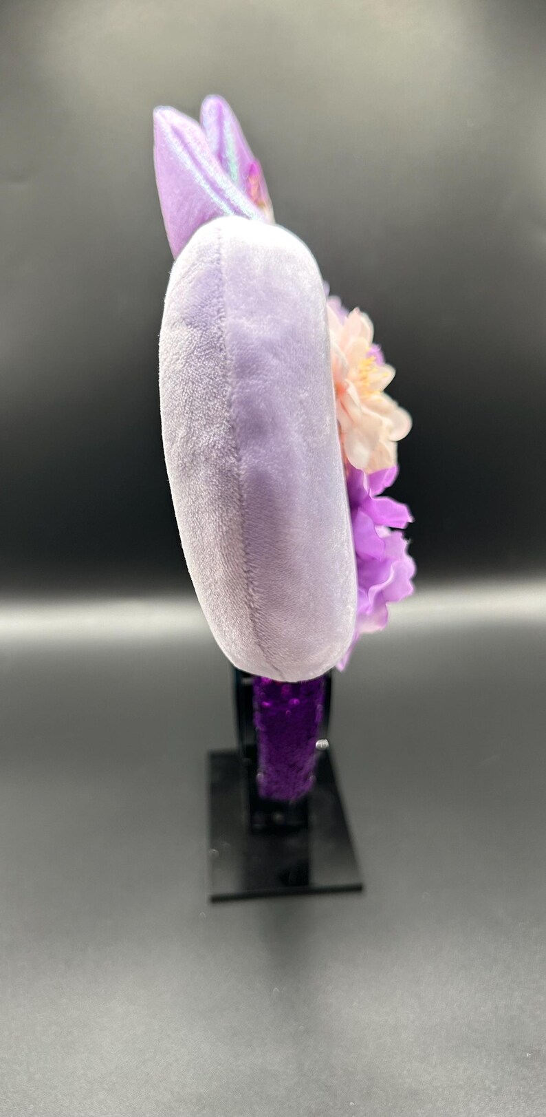 Puede incluir: Una vista lateral de una diadema con una oreja circular de felpa de color morado claro. La oreja est&aacute; decorada con flores artificiales en capas en tonos rosa p&aacute;lido y morado oscuro. La base de la diadema est&aacute; cubierta con un tejido texturizado de color morado oscuro.
