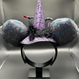 May include: A black headband with two large, round, textured ears. A purple witch hat with sequins and a black feather trim sits atop the headband. A small, red accent is attached to the hat. The ears have a shimmering, iridescent finish.