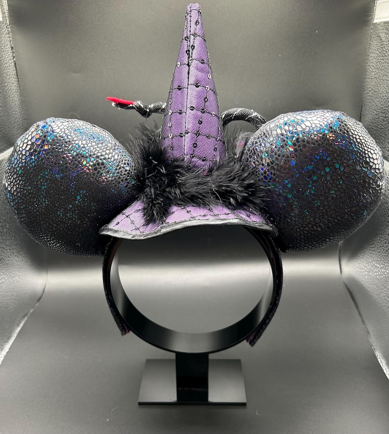 May include: A black headband with two large, round, textured ears. A purple witch hat with sequins and a black feather trim sits atop the headband. A small, red accent is attached to the hat. The ears have a shimmering, iridescent finish.