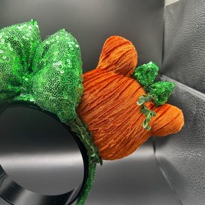 May include: A black headband with a green sequined bow and orange, textured Mickey Mouse ears. The ears have small green sequined accents. A festive accessory for themed events.