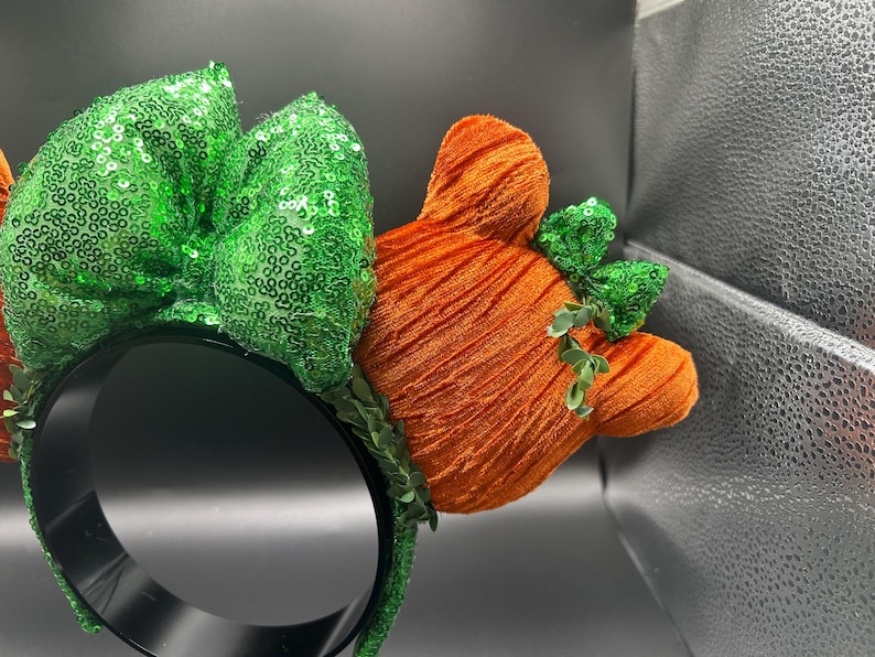May include: A black headband with a green sequined bow and orange, textured Mickey Mouse ears. The ears have small green sequined accents. A festive accessory for themed events.