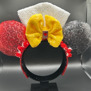 May include: A headband featuring two circular ears covered in red and black sequins. A central yellow bow sits between the ears, accented by a small playing card charm with a red heart. A white sequined fan-shaped piece rises behind the bow.