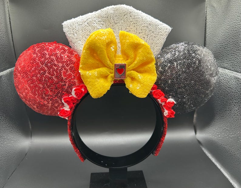 May include: A headband featuring two circular ears covered in red and black sequins. A central yellow bow sits between the ears, accented by a small playing card charm with a red heart. A white sequined fan-shaped piece rises behind the bow.