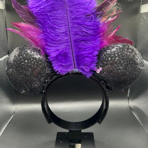 May include: A headband featuring two round black sequined ears and a large, upright purple ostrich feather plume. The base of the feathers is accented with smaller magenta feathers and a small, light pink three-circle icon on the right ear.