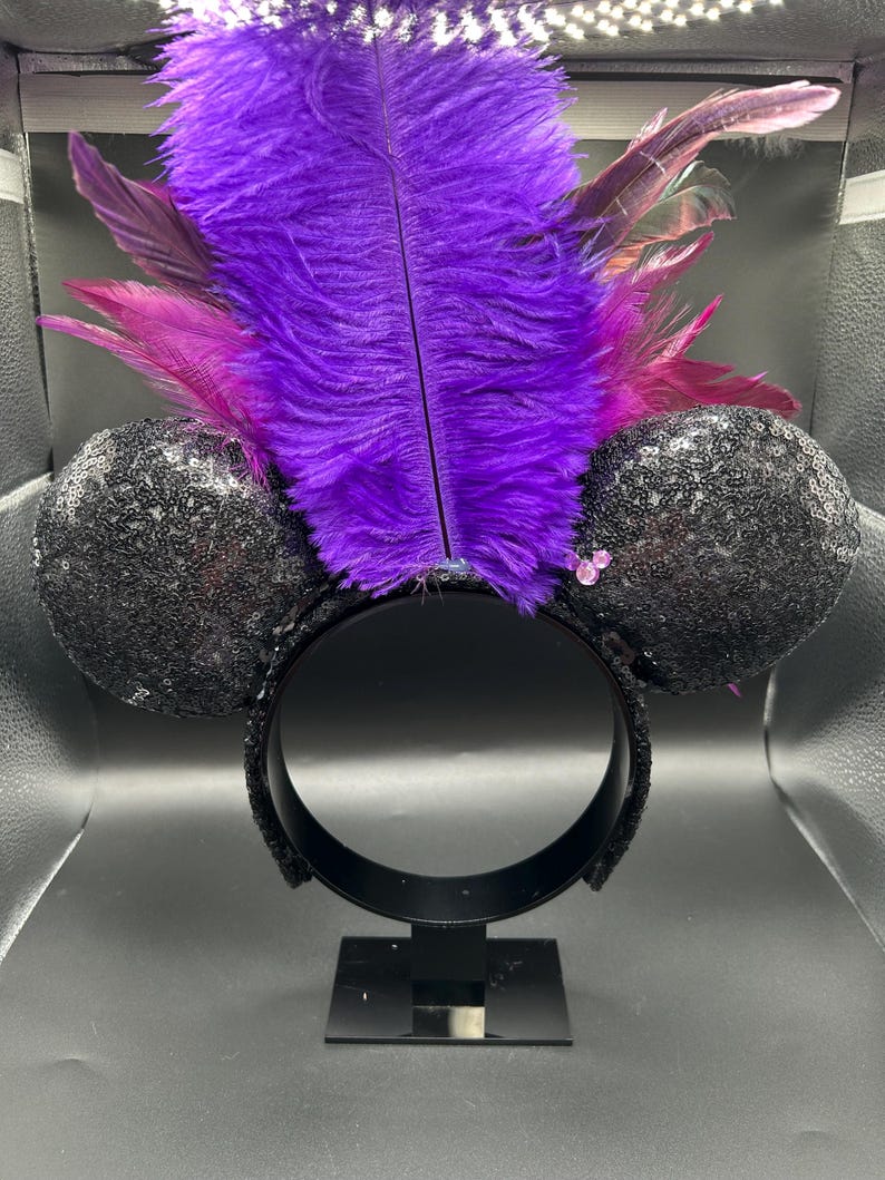 May include: A headband featuring two round black sequined ears and a large, upright purple ostrich feather plume. The base of the feathers is accented with smaller magenta feathers and a small, light pink three-circle icon on the right ear.
