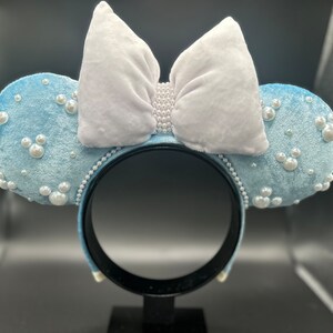 May include: Light blue velvet mouse ear headband with white bow and pearl accents. The ears are decorated with pearl clusters and Mickey Mouse silhouettes made of pearls. The headband is black.
