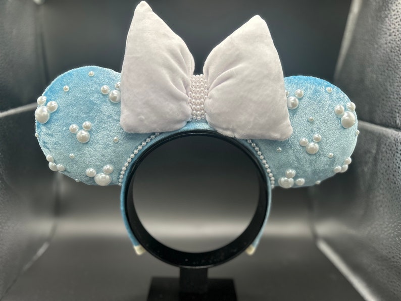 May include: Light blue velvet mouse ear headband with white bow and pearl accents. The ears are decorated with pearl clusters and Mickey Mouse silhouettes made of pearls. The headband is black.