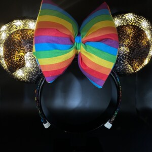 K&ouml;nnte beinhalten: Ein Paar schwarze M&auml;useohren mit einer regenbogenfarbenen Schleife und goldenen Glitzerohren. Die Ohren sind mit wei&szlig;en Lichtern beleuchtet.