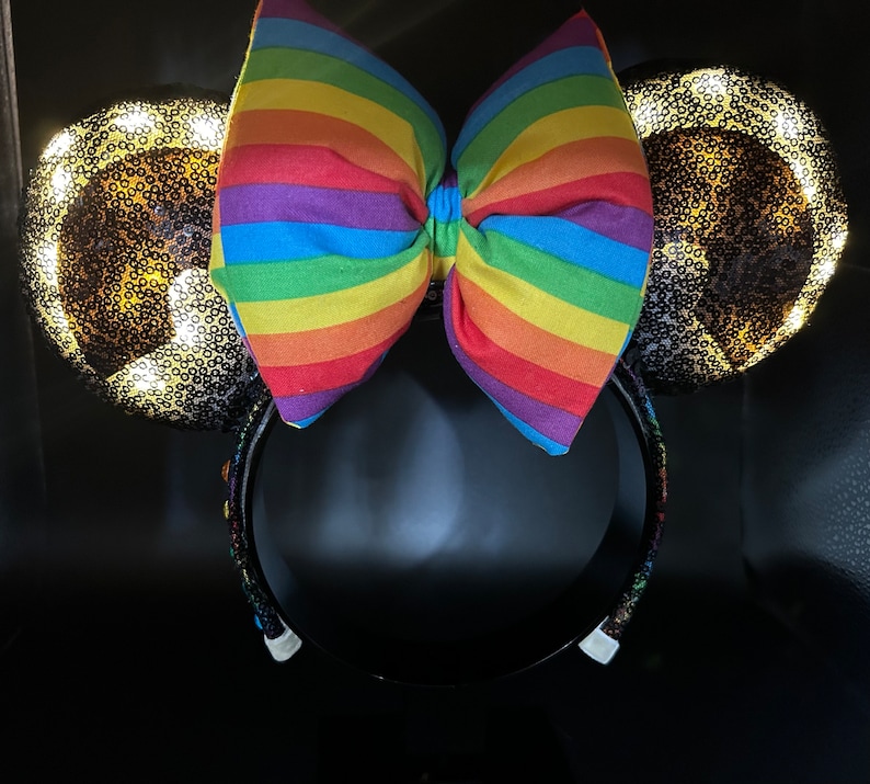 K&ouml;nnte beinhalten: Ein Paar schwarze M&auml;useohren mit einer regenbogenfarbenen Schleife und goldenen Glitzerohren. Die Ohren sind mit wei&szlig;en Lichtern beleuchtet.