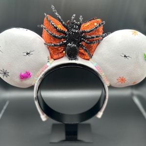 May include: Halloween-themed headband with white mouse ears decorated with black spiderwebs and spiders. A large orange sequined bow and a black glittery spider are centered on the headband. Pink and green embellishments add colour.
