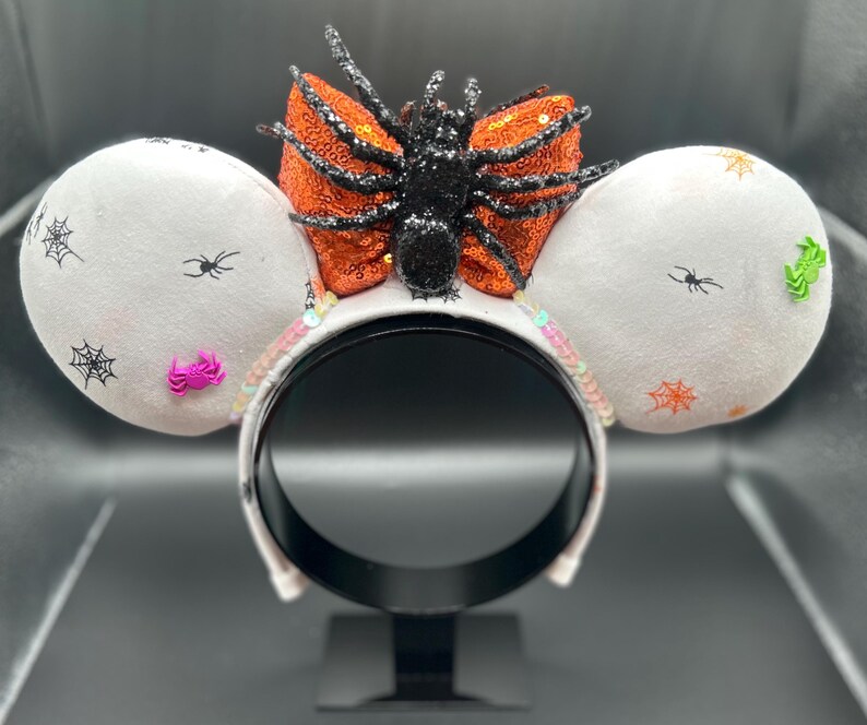 May include: Halloween-themed headband with white mouse ears decorated with black spiderwebs and spiders. A large orange sequined bow and a black glittery spider are centered on the headband. Pink and green embellishments add colour.