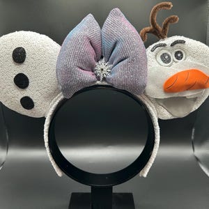 May include: A white headband with sequined ears and a bow. The left ear has three black dots, and the right ear is shaped like a snowman's head with a carrot nose and twig antlers. A silver snowflake is in the center of the bow.