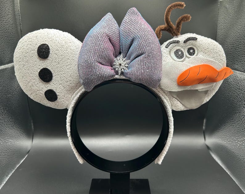 May include: A white headband with sequined ears and a bow. The left ear has three black dots, and the right ear is shaped like a snowman's head with a carrot nose and twig antlers. A silver snowflake is in the center of the bow.