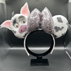 May include: A headband featuring two circular ears with a white and grey spotted pattern. The left ear depicts a pig character with pink inner ears and a glittery pink snout. A large, central bow covered in shimmering dark grey sequins sits between the ears.