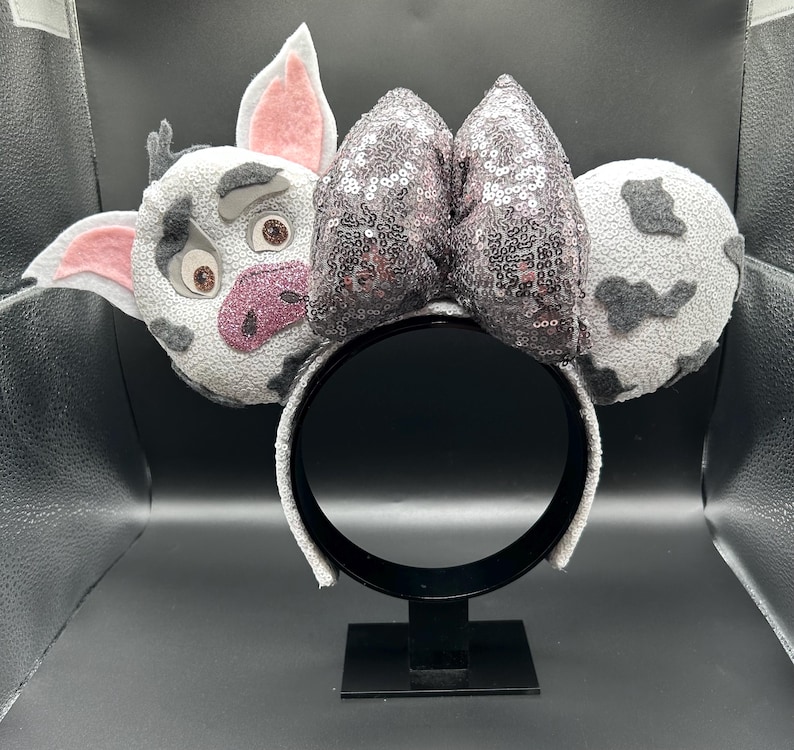 May include: A headband featuring two circular ears with a white and grey spotted pattern. The left ear depicts a pig character with pink inner ears and a glittery pink snout. A large, central bow covered in shimmering dark grey sequins sits between the ears.