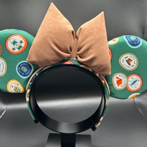 May include: Green fabric mouse ears with a brown bow. The ears are covered in a pattern of colourful patches with text like "Let's Go On A Hike" and "Happy Camper".